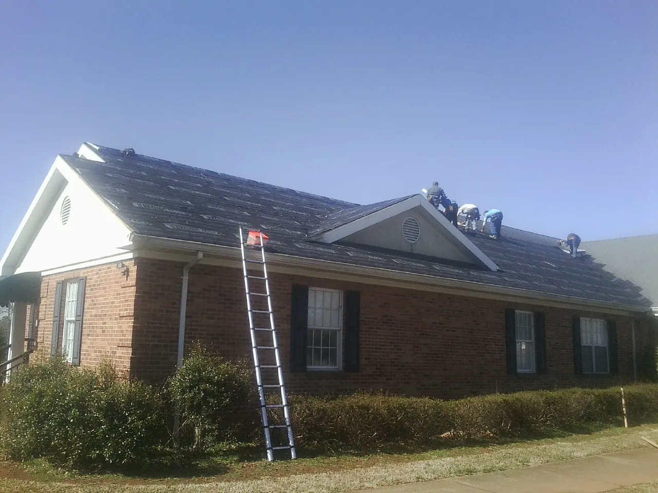 Crew tearing shingles off a brick ranch roof with a ladder set — Emergency Roof Repair work by our crew in Blue Rapids