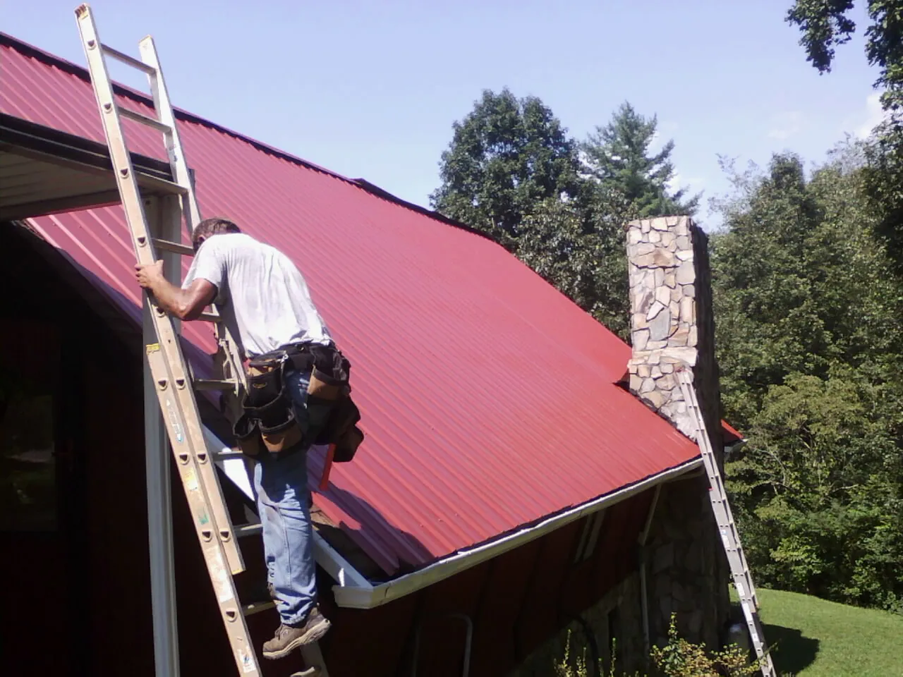 Roofer on a ladder beside a red metal roof and stone chimney — Soffit & Fascia Repair by Roofing Brothers in Blue Rapids